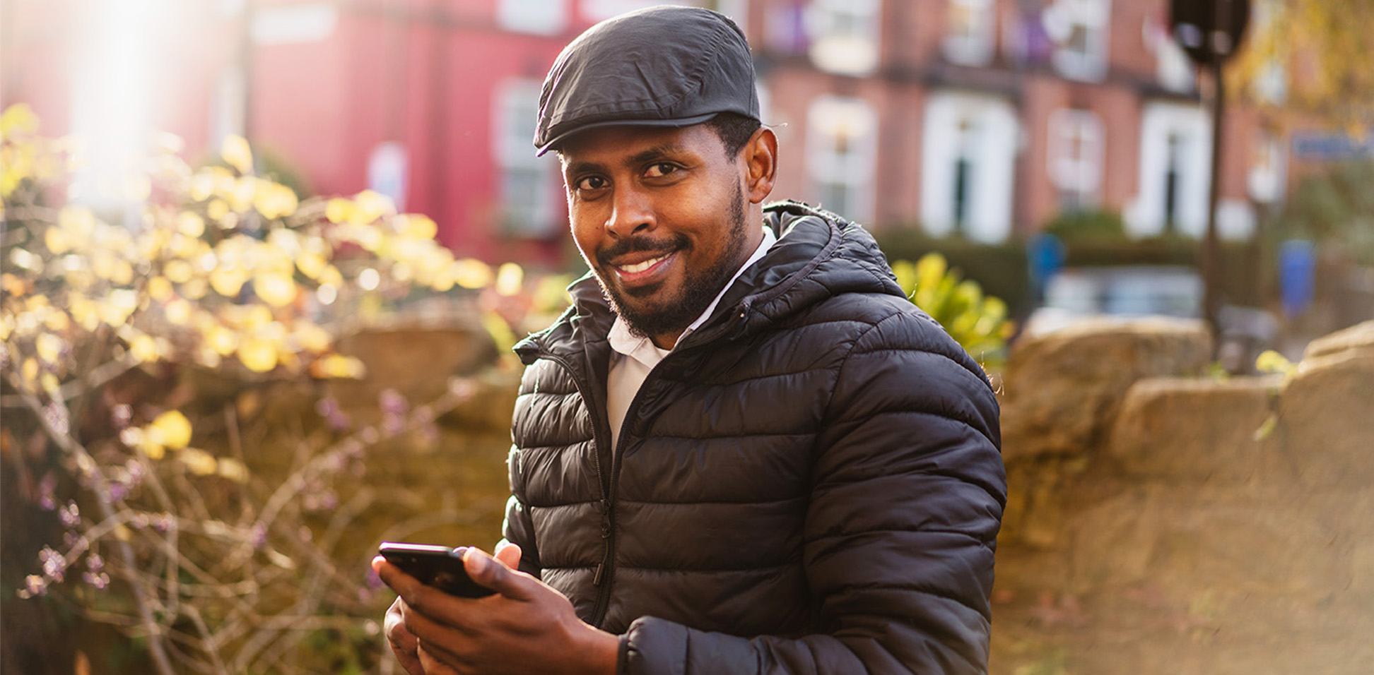 This photo shows a man smiling at the camera, stood outside holding a mobile phone.